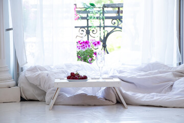 A small table with strawberries and a glass stand near the bed on the floor near the open balcony in the apartment, bedroom interior, breakfast in bed, romantic