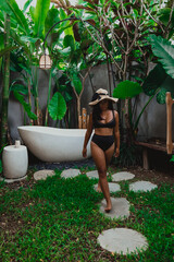 Modern outdoor bathroom with oval white ceramic bathtub decorated with green exotic plants and stones in luxury Bali villa with asian female model walking in black bikini swimsuit
