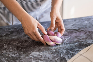 A woman bakes bread in the kitchen, braiding challah with a pigtail. The purple color of the challah is made from ebonized carrot powder.