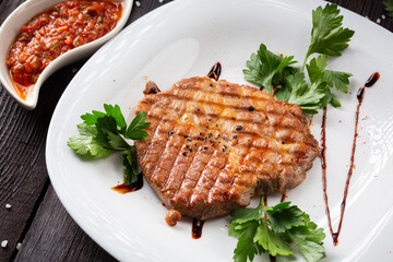 Pork meat steak under spicy salsa sauce with greens on a white plate. 