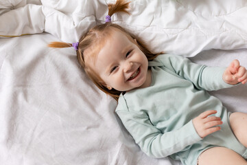 little girl in a light green bodysuit is lying in bed made with white bed linen