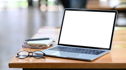 Computer laptop with white empty display, eyeglasses and notebooks on wooden office desk