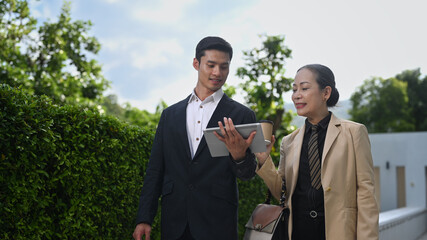 Smiling senior female investor consulting with young male financial adviser walking on the street in downtown of modern city