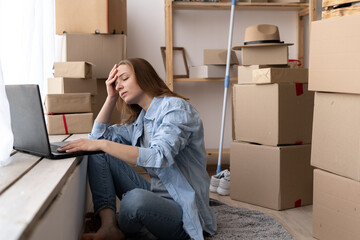 Young worried unhappy woman sitting on the floor near moving boxes using a laptop with hands on...