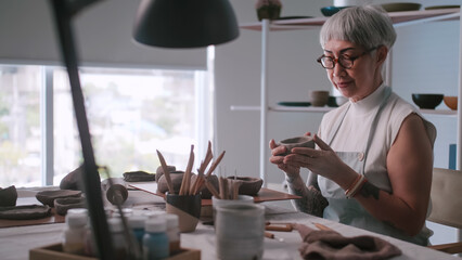 Asian elderly woman enjoying pottery work at home. A female ceramicist is making new pottery in a studio. - obrazy, fototapety, plakaty