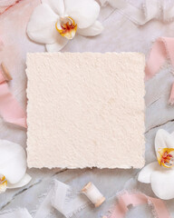 Handmade paper card near white orchid flowers and silk ribbons on marble, mockup