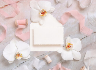 Handmade paper card and envelope near white orchid flowers and silk ribbons on marble, mockup