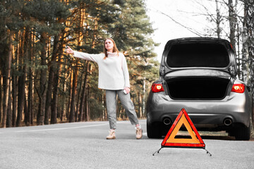 Red emergency triangle on the road with a blurry car in the background and a desperate woman catching passing cars waiting for help. Focus on emergency sign. Accident and broken car on the road.
