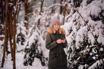 Young blonde girl in winter outfit stands in snowy park using smartphone