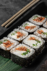 set of square maki rolls with fried salmon, curd cheese and green bamboo leaf in a black ceramic plate with chopstick on a dark gray textured background, side view, close-up