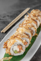 set of bonito rolls with grilled salmon, avocado, tuna shavings and green bamboo leaf in a white ceramic plate with chopstick on a dark gray textured background, side view, close-up