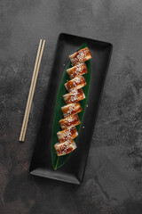 set of philadelphia rolls with eel, smoked salmon, avocado and green bamboo leaf in a black ceramic plate with chopstick on a dark gray textured background, top view