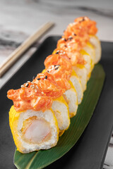 set of yellow maki takuan rolls with tiger shrimp, avocado, salmon and green bamboo leaf in a black ceramic plate with chopstick on a bright white textured marble background, side view, close-up