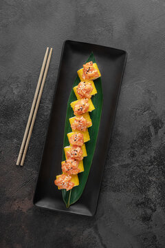 set of yellow maki takuan rolls with tiger shrimp, avocado, salmon and green bamboo leaf in a black ceramic plate with chopstick on a dark gray textured background, top view