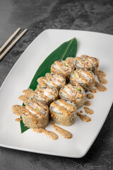 set of fried rolls with tiger shrimp, avocado, sesame sauce and green bamboo leaf in a white ceramic plate with chopstick on a dark gray textured background, side view