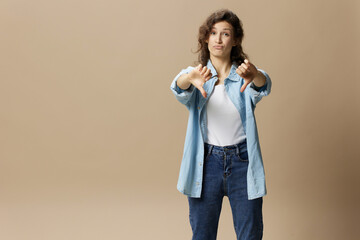 Unhappy curly beautiful female in jeans casual shirt show thumbs down gesture Dislike at camera posing isolated on over beige pastel background. People Lifestyle emotions concept. Copy space