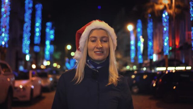 A beautiful woman in a Santa Claus hat is walking on a winter night. The street is decorated with garlands of flowers along the alley, decorated with lights on New Year's Eve or Christmas.