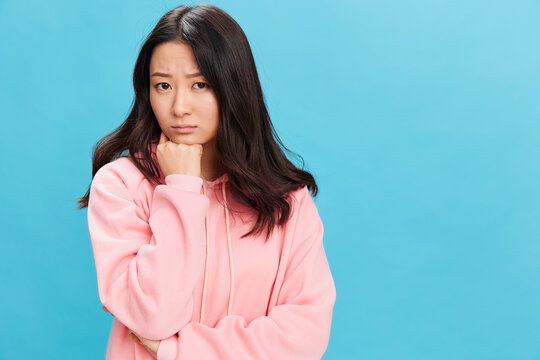 Upset sad cute Asian student young lady in pink hoodie sweatshirt folds hands posing isolated on over blue studio background. The best offer for ad. People Emotions for Everyday concept
