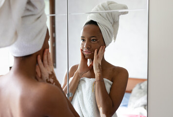Portrait of woman with vitiligo wearing towel applying moisturizing cream on the face, looking at...