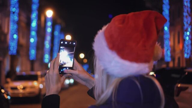 A smiling woman in a Santa Claus hat taking a photo of the night city with her smartphone on Christmas eve. A happy woman takes pictures on her mobile phone on the eve of the Christmas holidays.