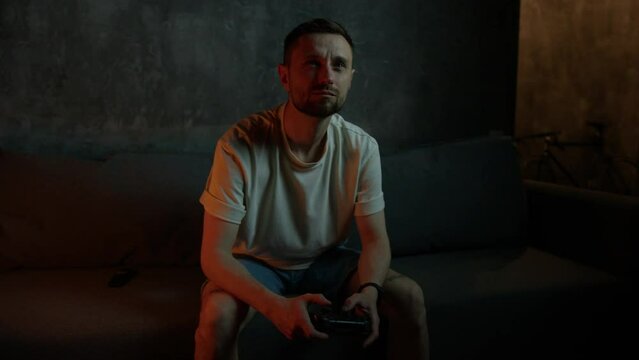 A male gamer is sitting at home on the couch and holding a joystick in his hands. Video games at home