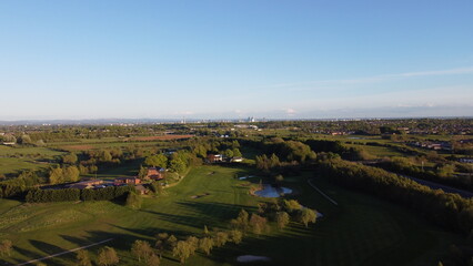 Aerial view looking down onto a golf course surrounded by green fields with the Manchester skyline in the distance. 