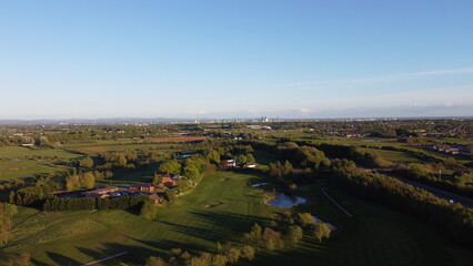 Aerial view looking down onto a golf course surrounded by green fields with the Manchester skyline in the distance. 