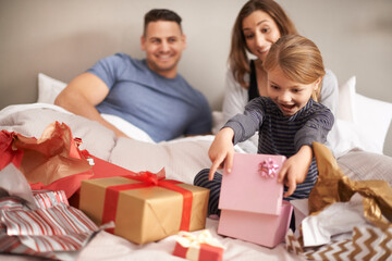 Oh wow, thanks mom and dad. Shot of a little girl opening presents in bed as her parents watch.