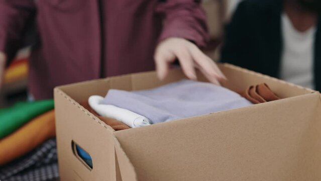 Caucasian woman sorting and putting clothes at the box while working at the reuse center with her colleague. Second hand and humanitarian aid concept. Cropped view