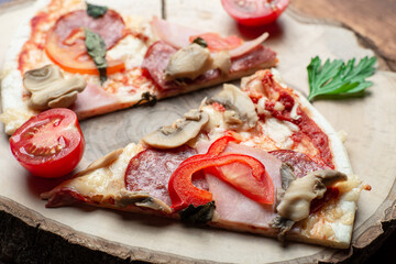 Pizza with ham and bacon, mushrooms and tomatoes on a slice of wood on a brown wooden background.