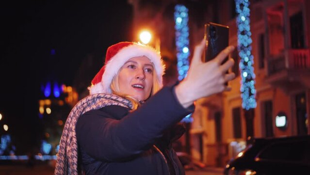 A young beautiful girl on Christmas walks around the city at night and takes a selfie. Christmas lights.She is wearing a Santa hat and taking pictures on her smartphone for social media.