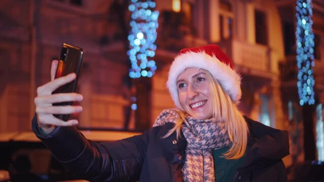 A young beautiful girl on Christmas walks around the city at night and takes a selfie. Christmas lights.She is wearing a Santa hat and taking pictures on her smartphone for social media.
