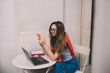 Young Caucasian student sitting with smartphone front open laptop computer while learning online. Millennial female browsing mobile internet via application. Freelancer using technology for work