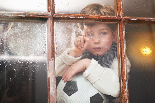 Waiting for some sunshine. A young boy sitting by the window and looking bored while it rains outside.