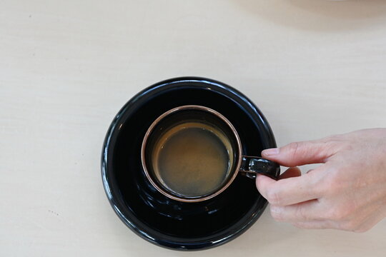 Left Hand Of Asian Woman (white Skin) Holding A Black Espresso Cup Served On A Wooden Table, Lying Down, Top View.
