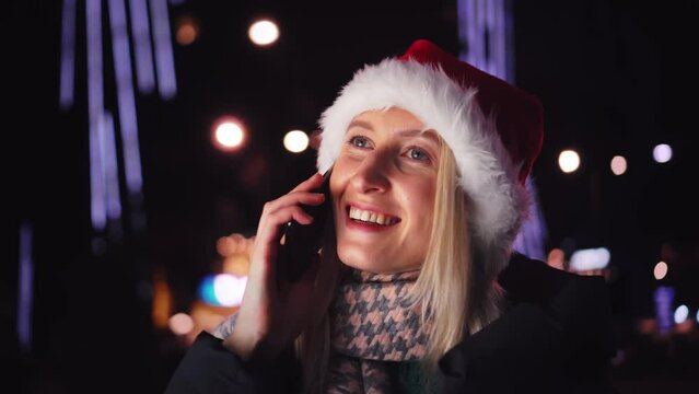 Close-up of a young beautiful woman in winter clothes, emotionally and wonderfully talking on mobile phone in a Santa Claus hat against background of a night city and lights. She is happy and smiling.