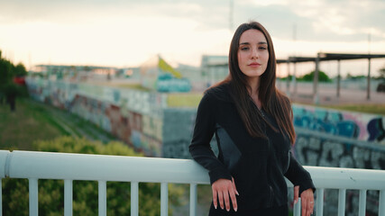 Portrait of cute girl with freckles and loose brown hair wearing black sports hoodie looks at the camera and walks on the bridge of the modern city background