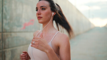 Young athletic woman with long ponytail wearing beige sports top in wired headphones, runs along...