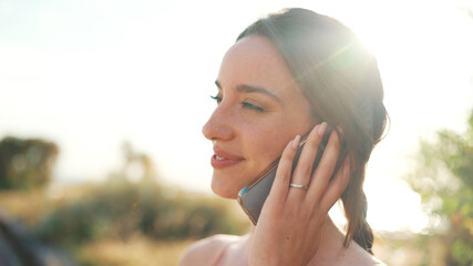 Lovely smiling girl with plait wearing beige sports top talking on smartphone sitting on bench. Young woman communicate in mobile phone. Back light