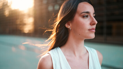 Close-up portrait of young woman with freckles and dark loose hair and long eyelashes wearing white top looking straight at the camera. Beautiful girl on modern city background