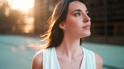 Close-up portrait of young woman with freckles and dark loose hair and long eyelashes wearing white top looking straight at the camera. Beautiful girl on modern city background