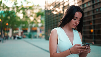 Beautiful woman with freckles and dark loose hair wearing white top is walking down the street with smartphone in her hands. Girl uses mobile phone on modern city background