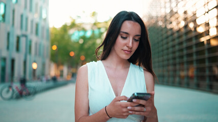 Beautiful woman with freckles and dark loose hair wearing white top is walking down the street with smartphone in her hands. Girl uses mobile phone on modern city background