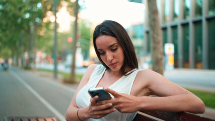Beautiful woman with freckles and dark loose hair wearing white top sits on bench with phone in her hands. Cute smiling girl watching photo, video on mobile phone modern city background