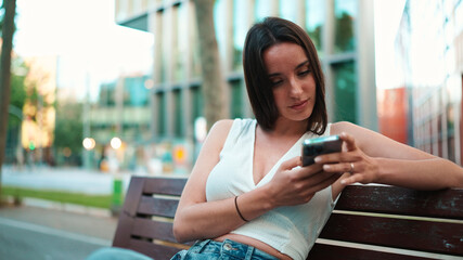 Beautiful woman with freckles and dark loose hair wearing white top sits on bench with phone in her hands. Cute smiling girl watching photo, video on mobile phone modern city background