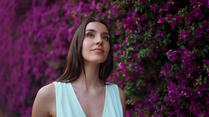 Beautiful woman with freckles and dark loose hair wearing white top walks along wall blooming with purple flowers. Cute girl looking at purple flowers