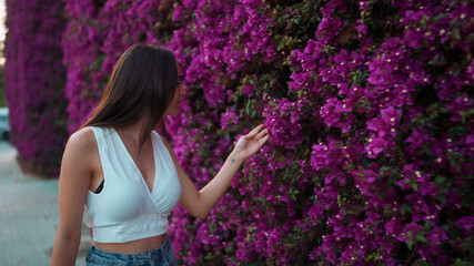 Beautiful woman with freckles and dark loose hair wearing white top walks along wall blooming with purple flowers. Cute girl looking at purple flowers