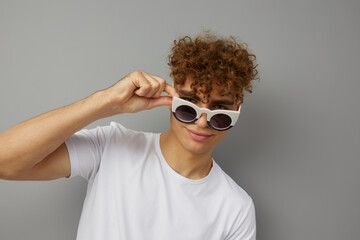 a young man stands in a casual T-shirt on an isolated gray background with sunglasses on and holding them with his hand looks at the camera