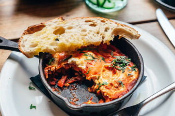 Half eaten freshly made shakshuka with spiced tomato, red pepper, feta, egg, coriander and pieces of homemade sourdough, served in iron pan, healthy vegetarian breakfast