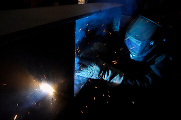 A welder in protective overalls and a welding mask welds metal structures by welding in a...